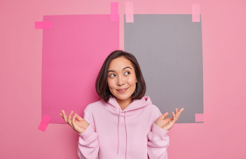 Uncertain hesitant Asian woman spreads palms feels unaware thinks about choice has no idea being clueless stands questioned wears casual hoodie poses against blank empty background. So what.